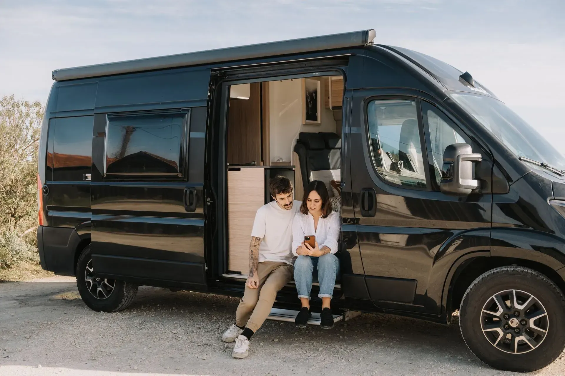 Black camper van with side door open, two people sitting at the entrance in a desert setting.