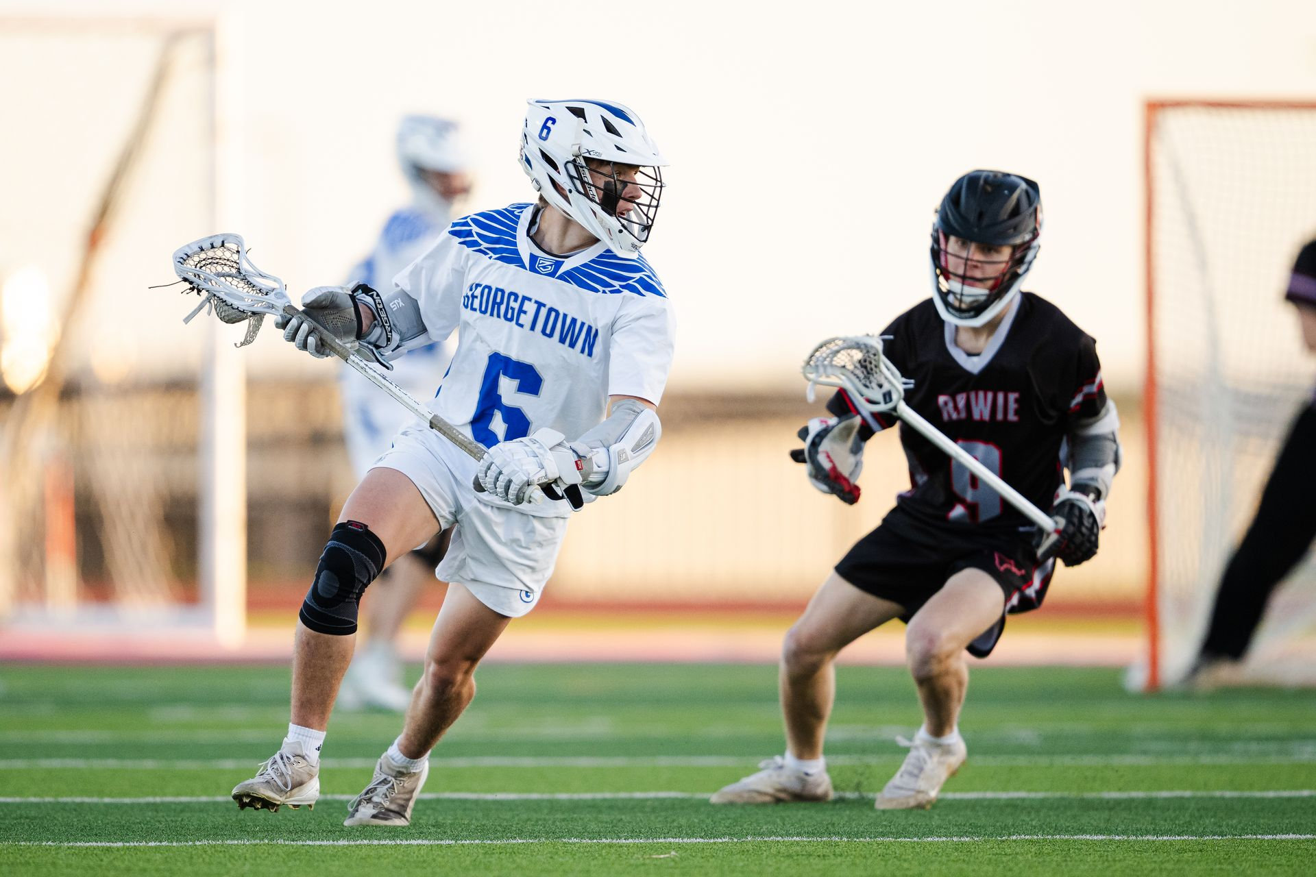 Two lacrosse players sprint after the ball, one in white and blue, one in black, on a field with a goal in view.