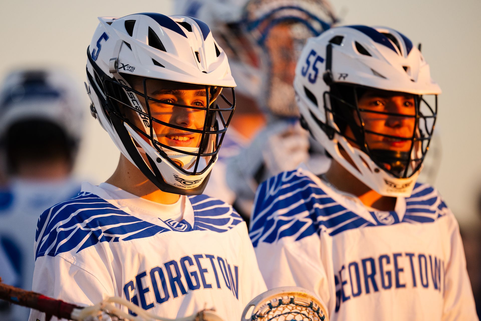 Two Georgetown lacrosse players in white uniforms and helmets standing on the field