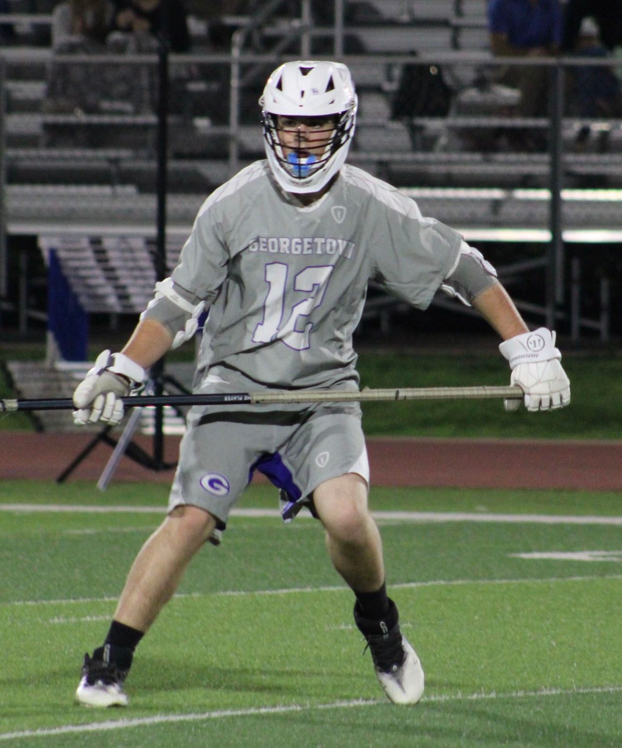 Lacrosse player in gray uniform running on a field at night, holding a stick horizontally.