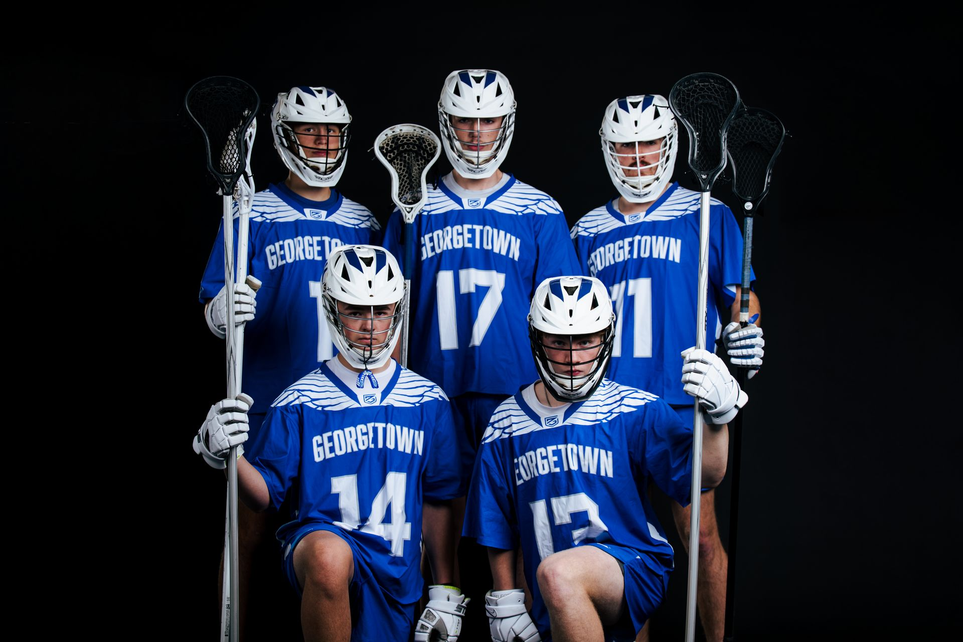 Five lacrosse players in blue jerseys posing with sticks against a black background