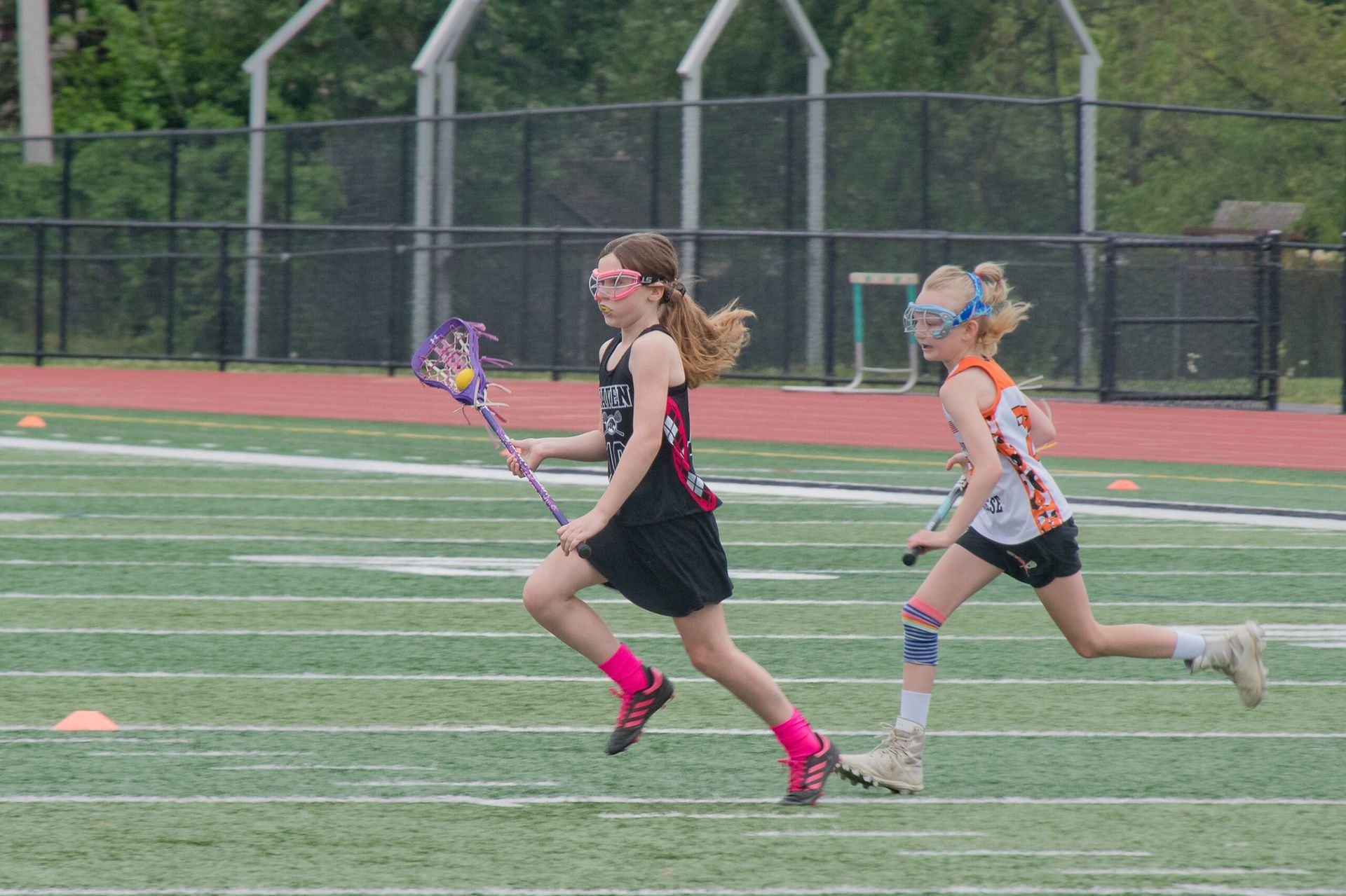 Two lacrosse players sprinting on a field, one in black with pink shoes and one in orange