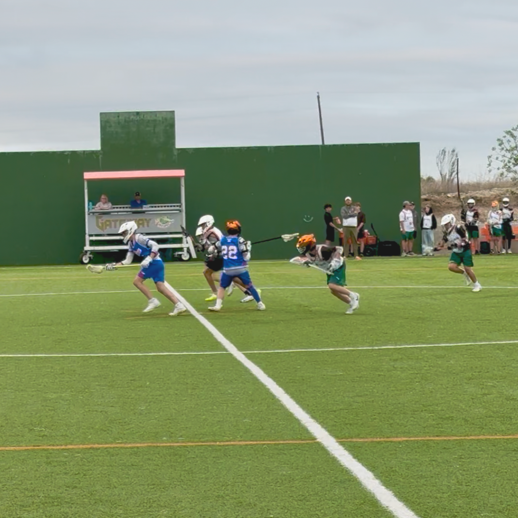 Lacrosse players in blue and black sprint across a green field during a game.