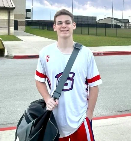 Smiling student in white sports jersey with red trim, holding a black duffel bag outdoors near a school track.