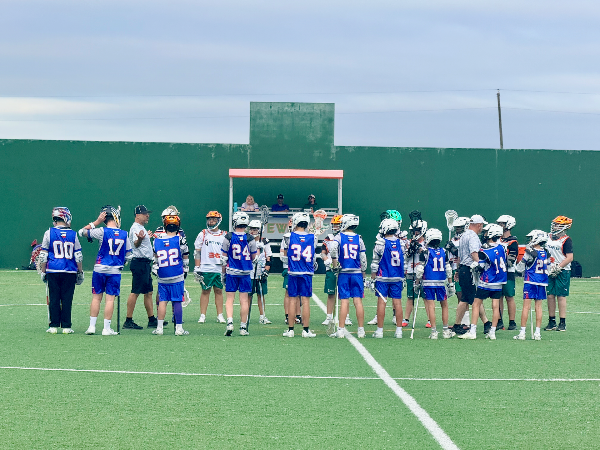 Youth lacrosse team in blue uniforms gathered on a field near the goal during a game timeout