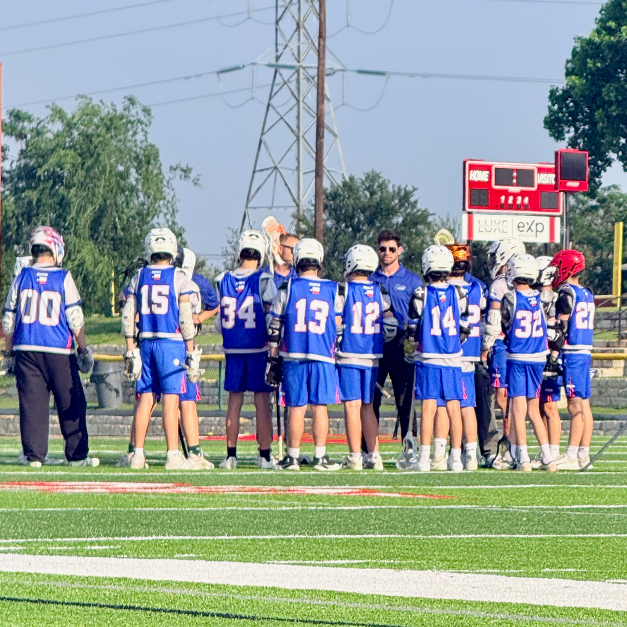 Youth lacrosse team in blue uniforms huddles on a field near a scoreboard.
