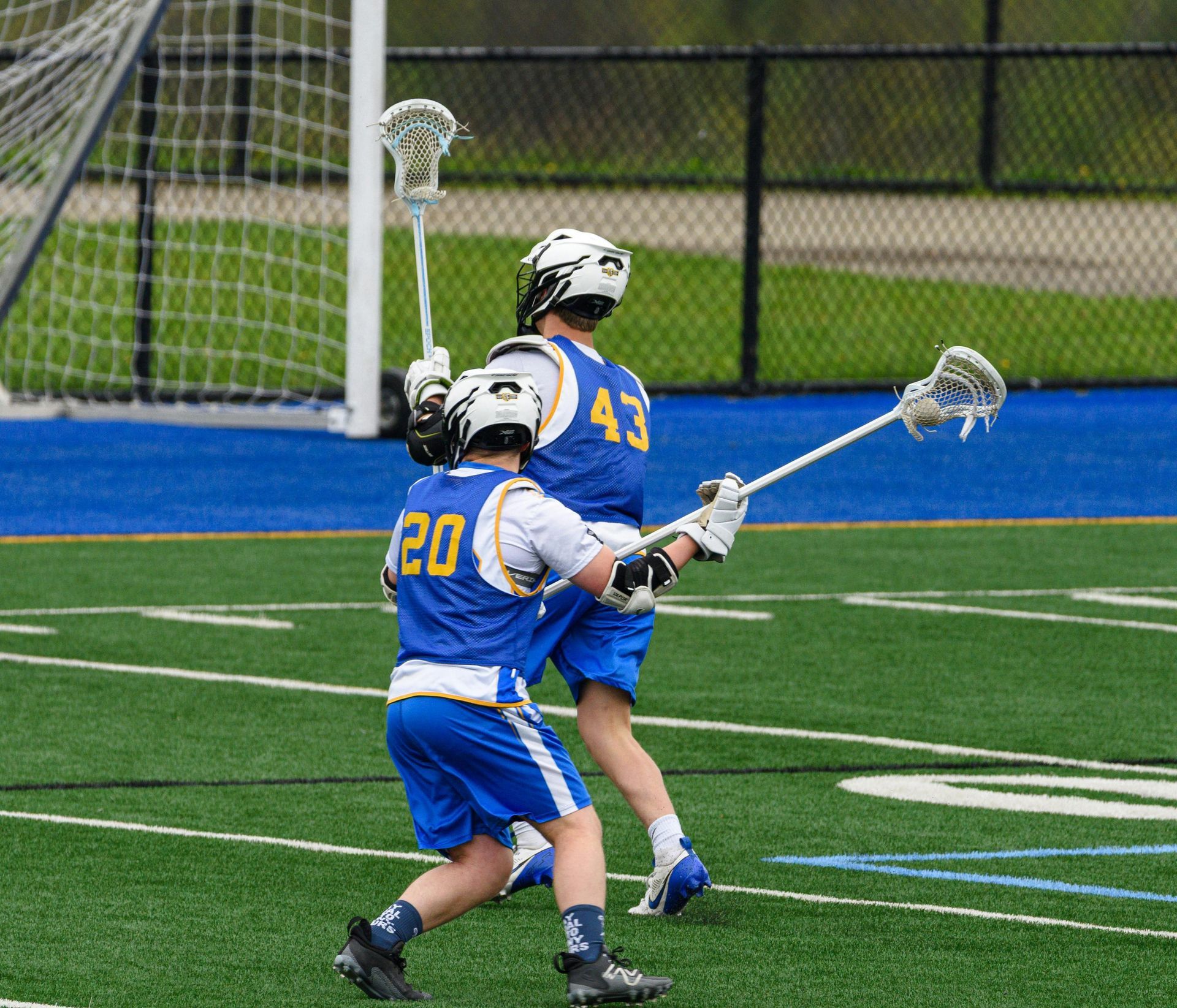 Two lacrosse players in blue uniforms battle for the ball near the goal on a field.