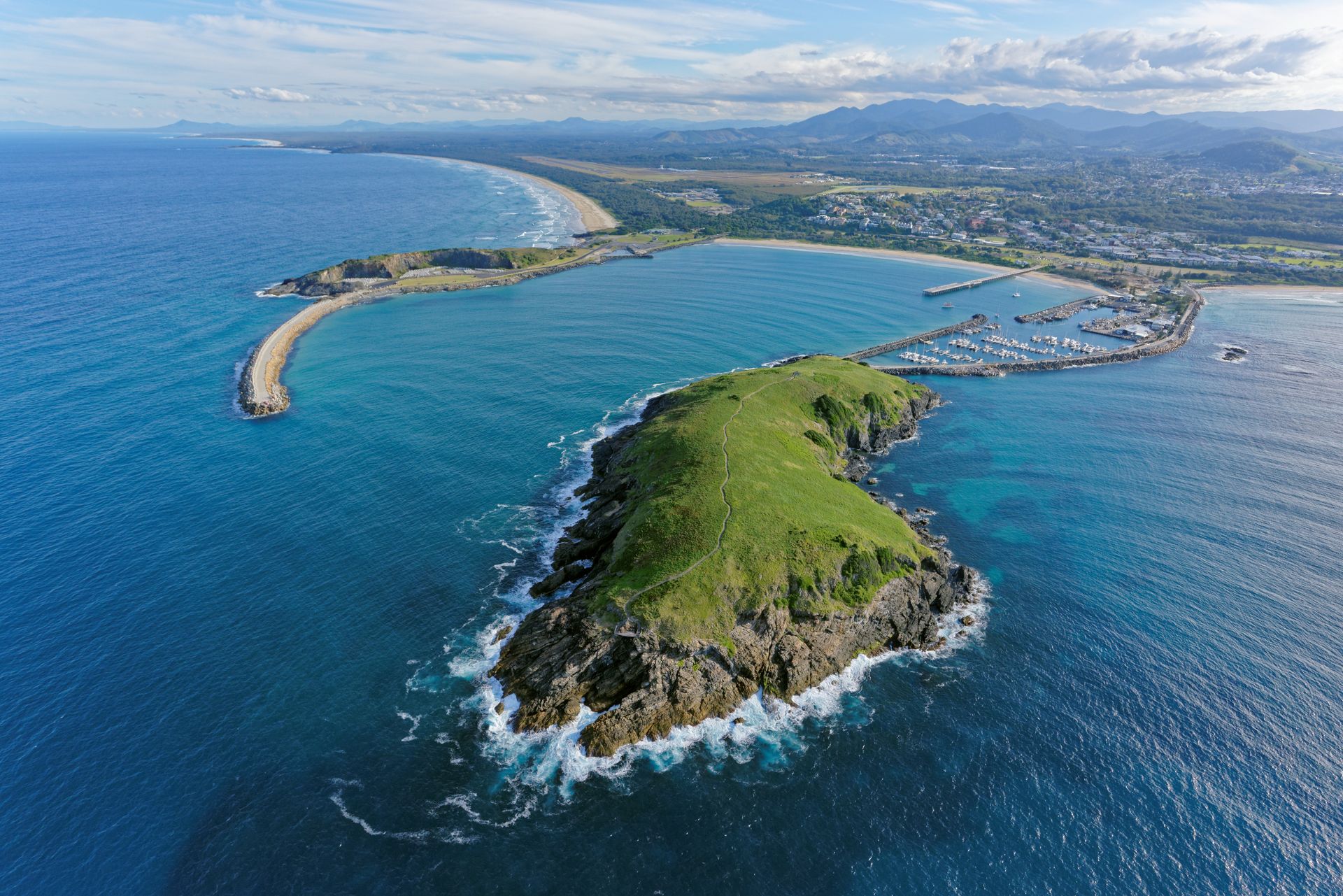 Aerial view of a green island beside a curved sandy beach and blue ocean under a partly cloudy sky