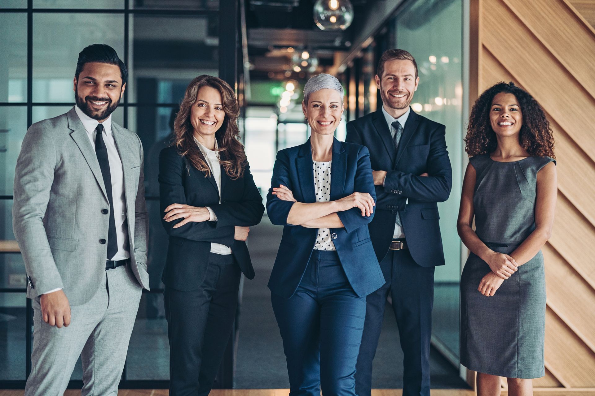Five professionals standing together in a modern office, arms crossed and smiling at the camera.