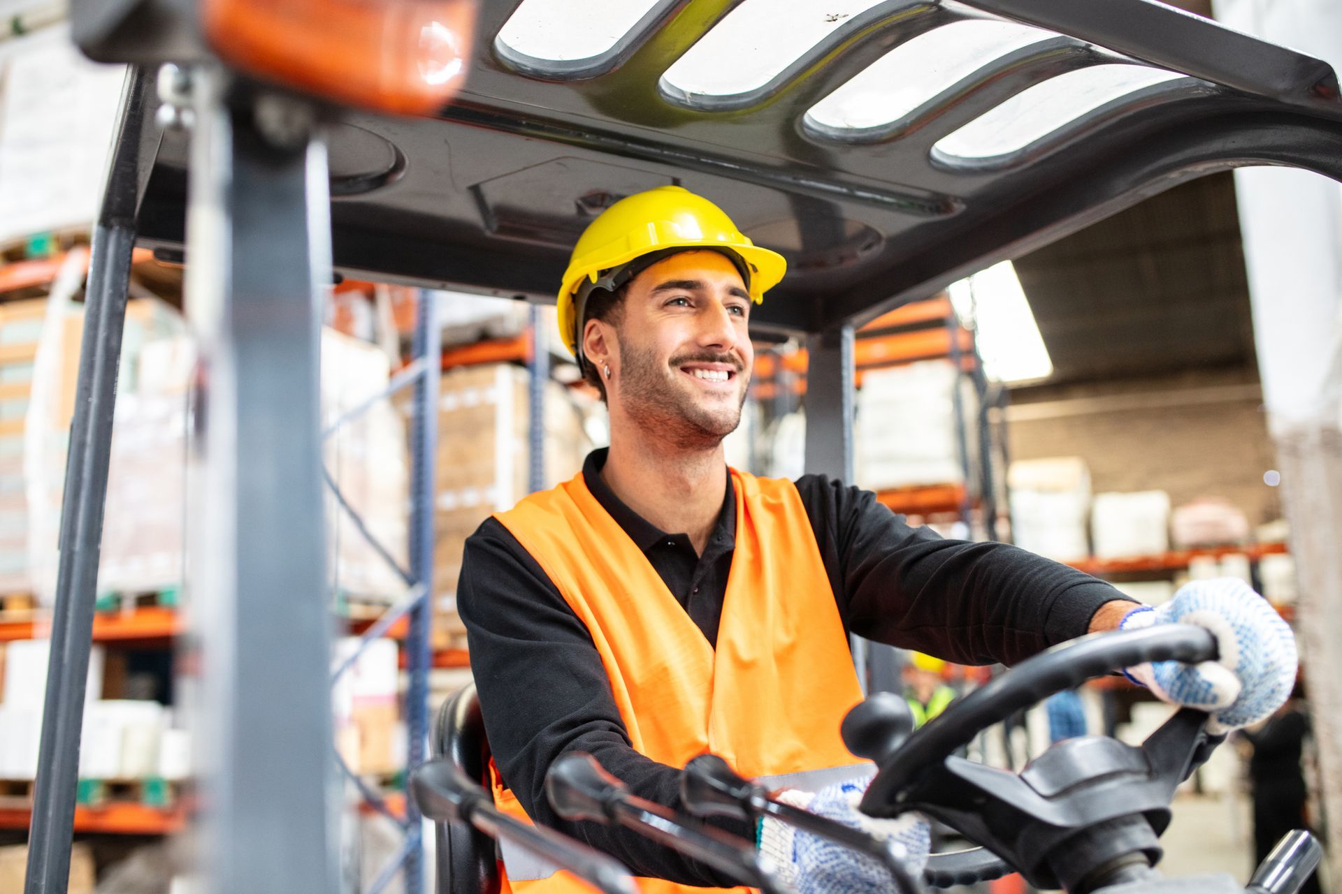 Warehouse worker in yellow hard hat and orange vest driving a forklift, smiling in an industrial setting