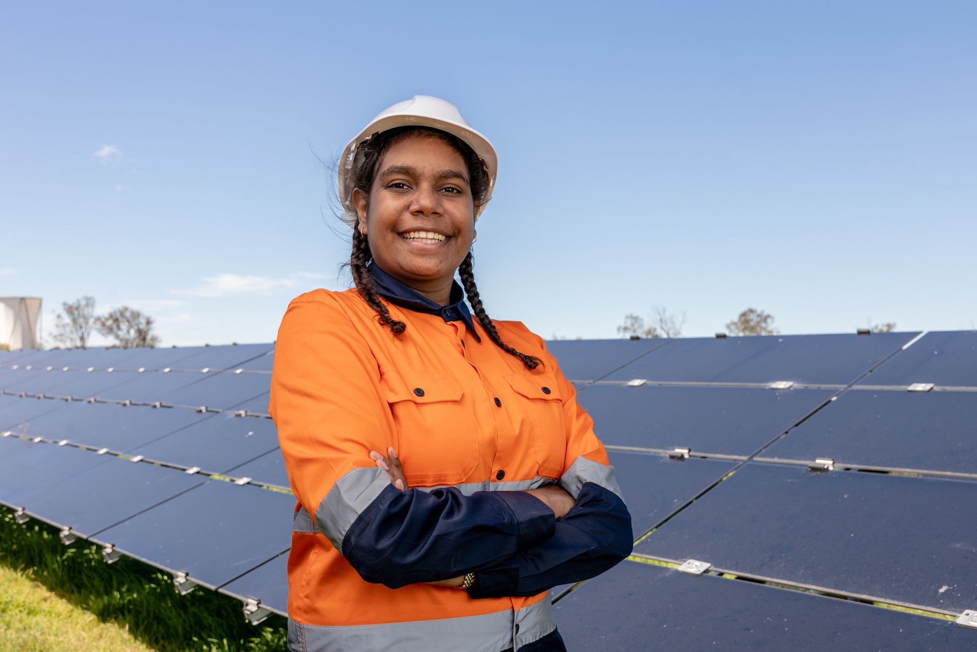 Person in orange safety jacket and white hard hat standing before solar panels outdoors, arms crossed, smiling