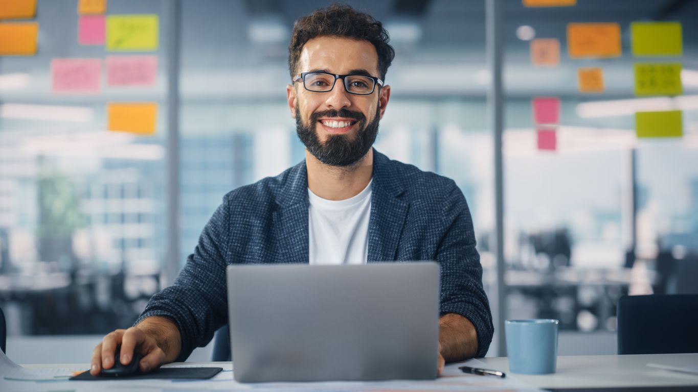 Smiling person at a desk with a laptop in a bright office, with sticky notes on the glass wall behind.