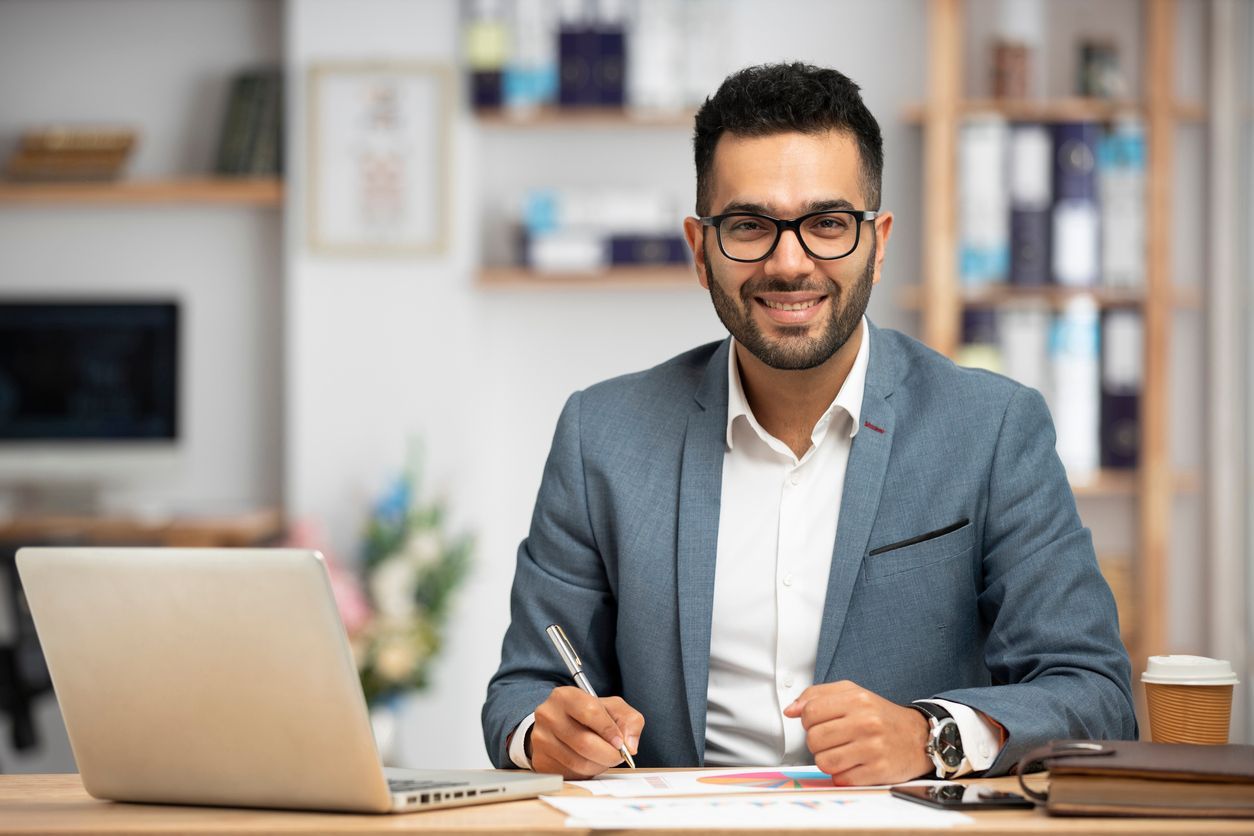 Smiling man in a gray blazer working at a desk with a laptop and notebook in a bright office