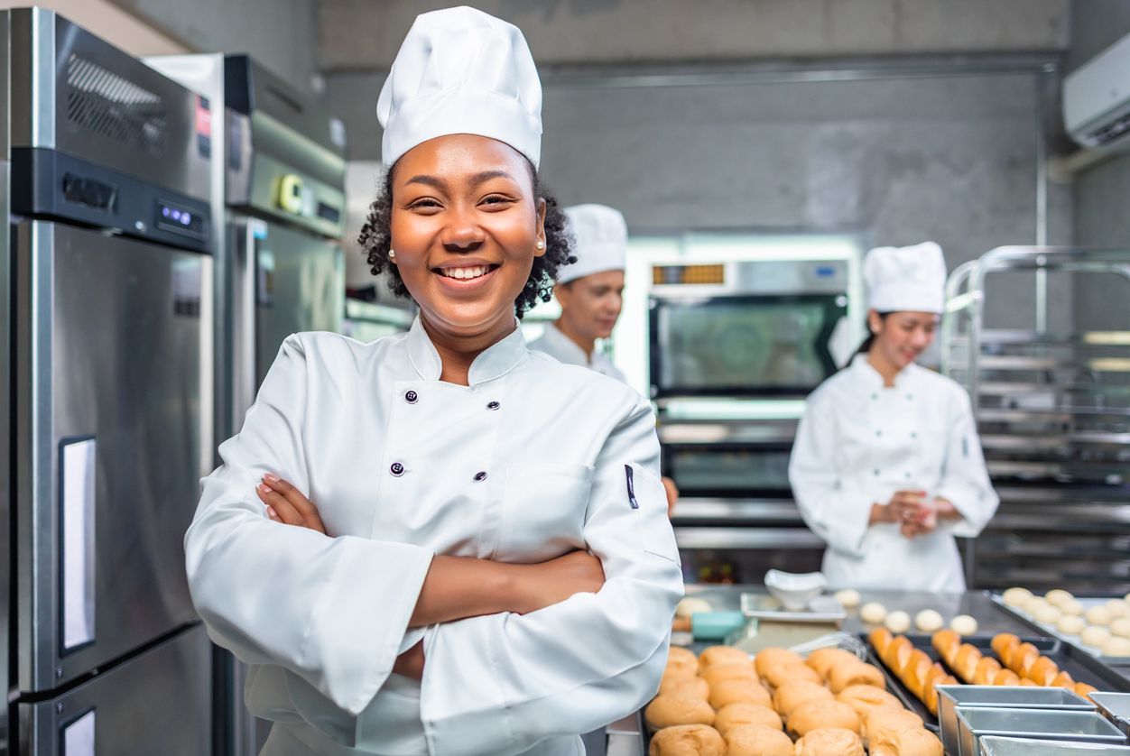 Chef smiling in a commercial kitchen, standing with arms crossed beside trays of fresh baked goods.