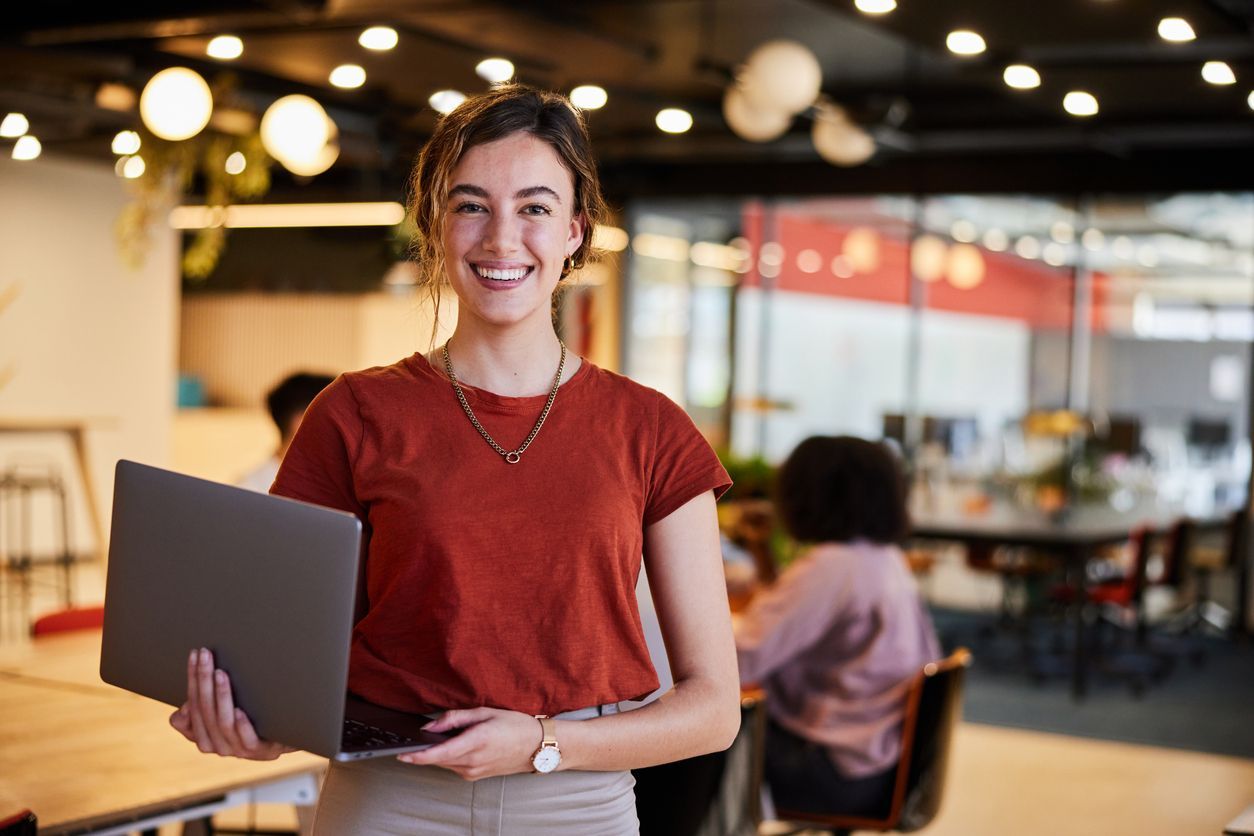 Smiling person holding a laptop in a modern office, with warm lights and coworkers in the background