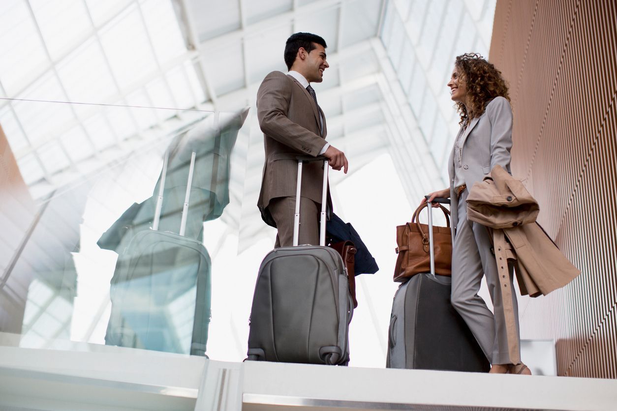 Man and woman in business attire, standing with luggage in a modern airport, smiling and talking.