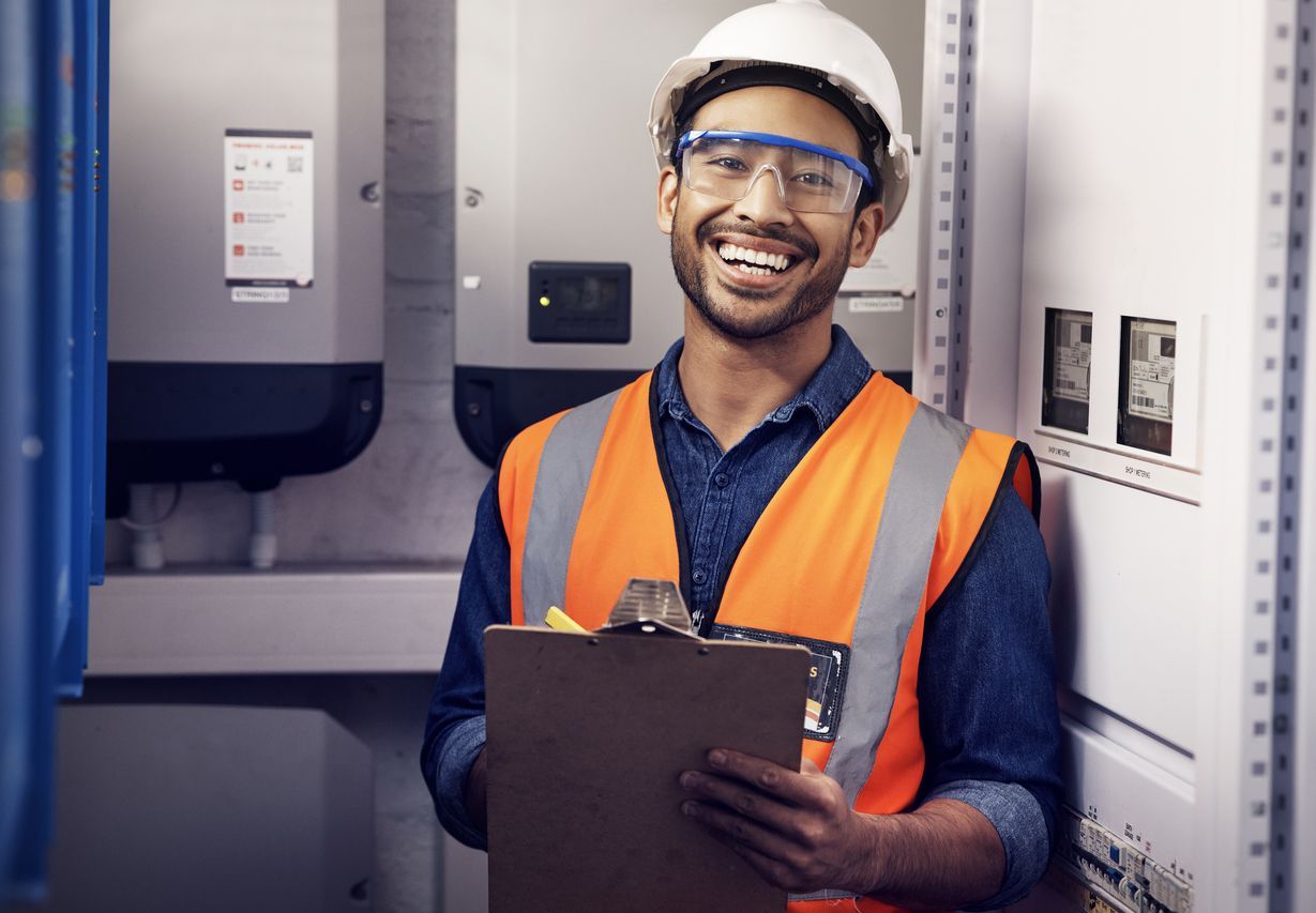 Smiling technician in orange safety vest and hard hat holding a clipboard in an electrical room