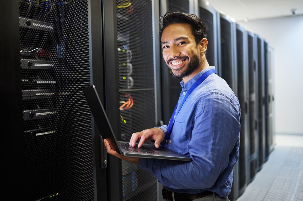 Technician smiling while using a laptop in a server room beside racks of network equipment
