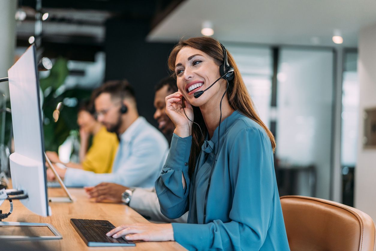 Call center agent in blue blouse wearing headset, smiling at a desk in a bright office