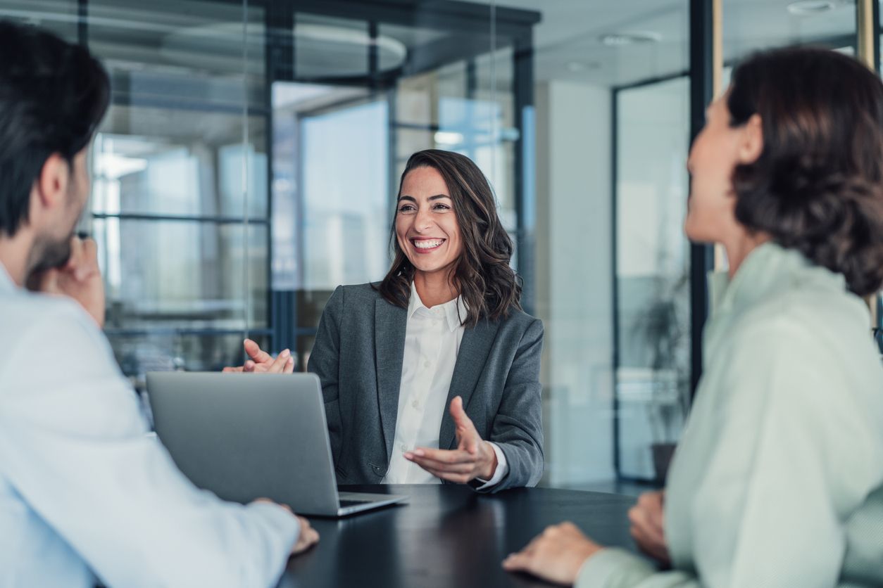 Three coworkers in a glass office meeting, one smiling and speaking across a laptop