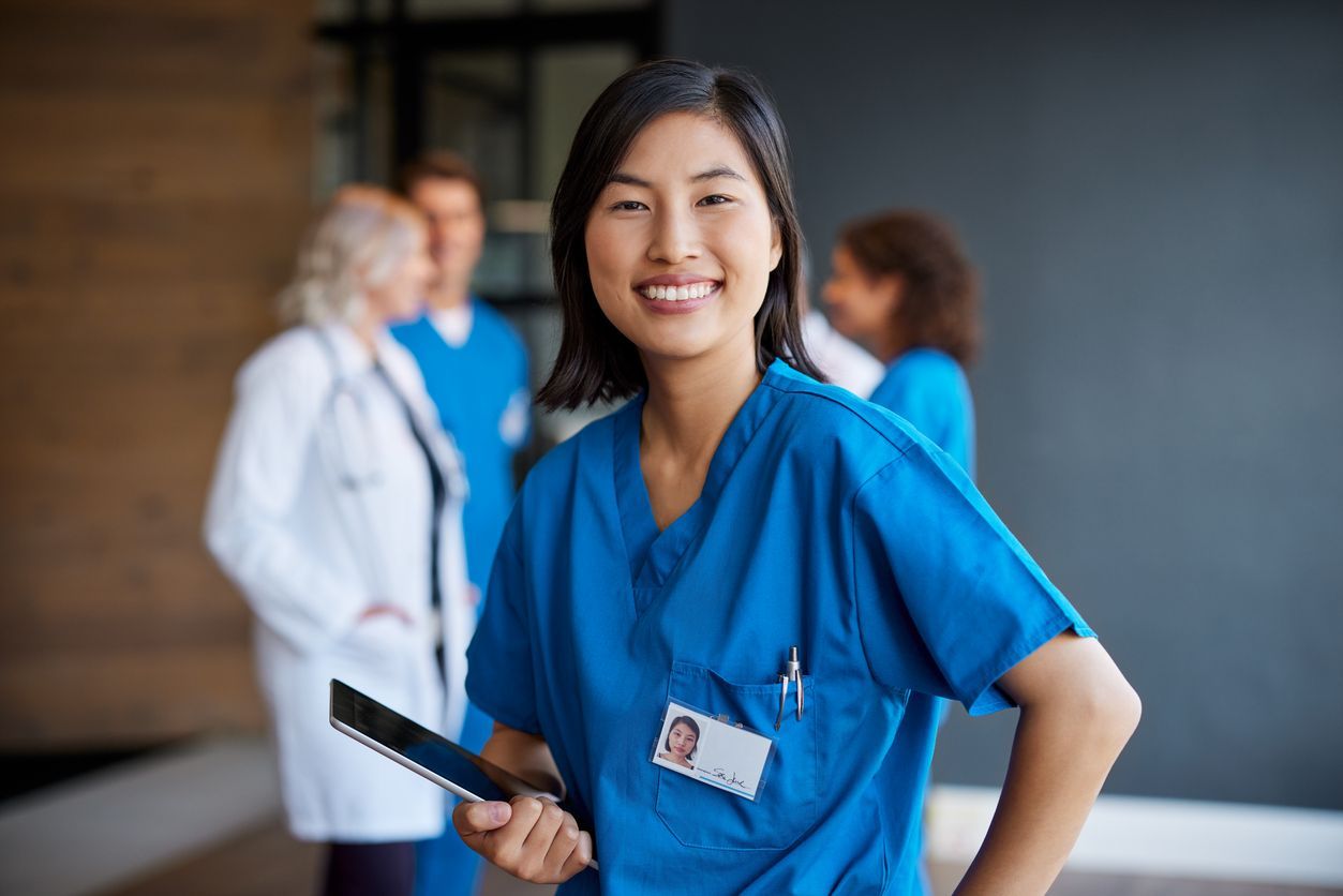 Smiling healthcare worker in blue scrubs holding a tablet, with colleagues blurred in the background.