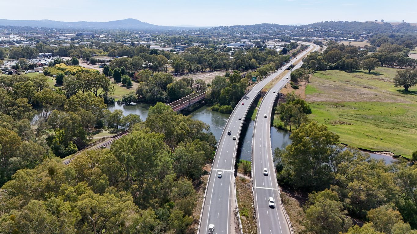 Aerial view of a highway crossing a river through green trees and open fields, with city buildings in the distance.