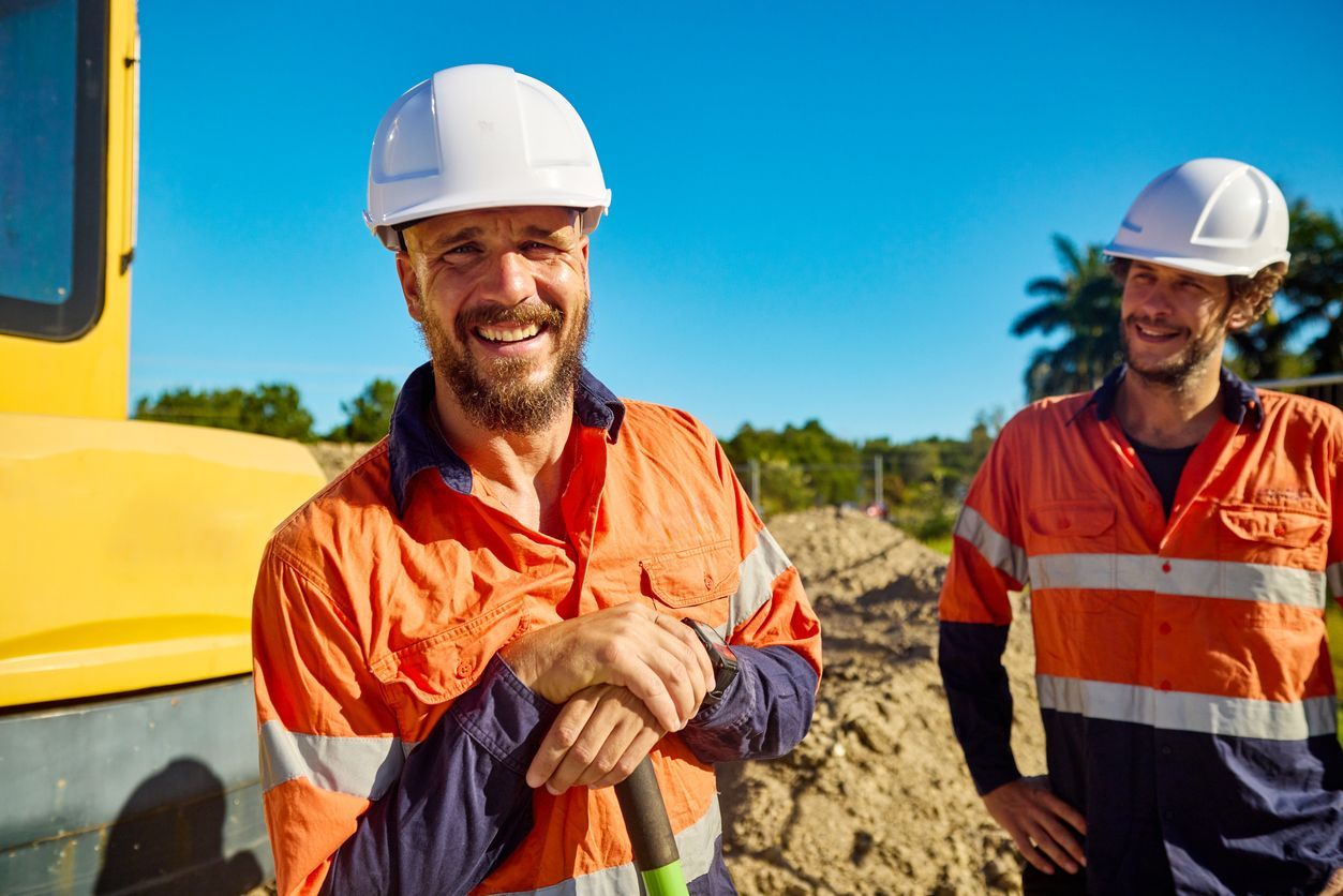 Two construction workers in white hard hats and orange safety gear standing at a construction site with a yellow machine