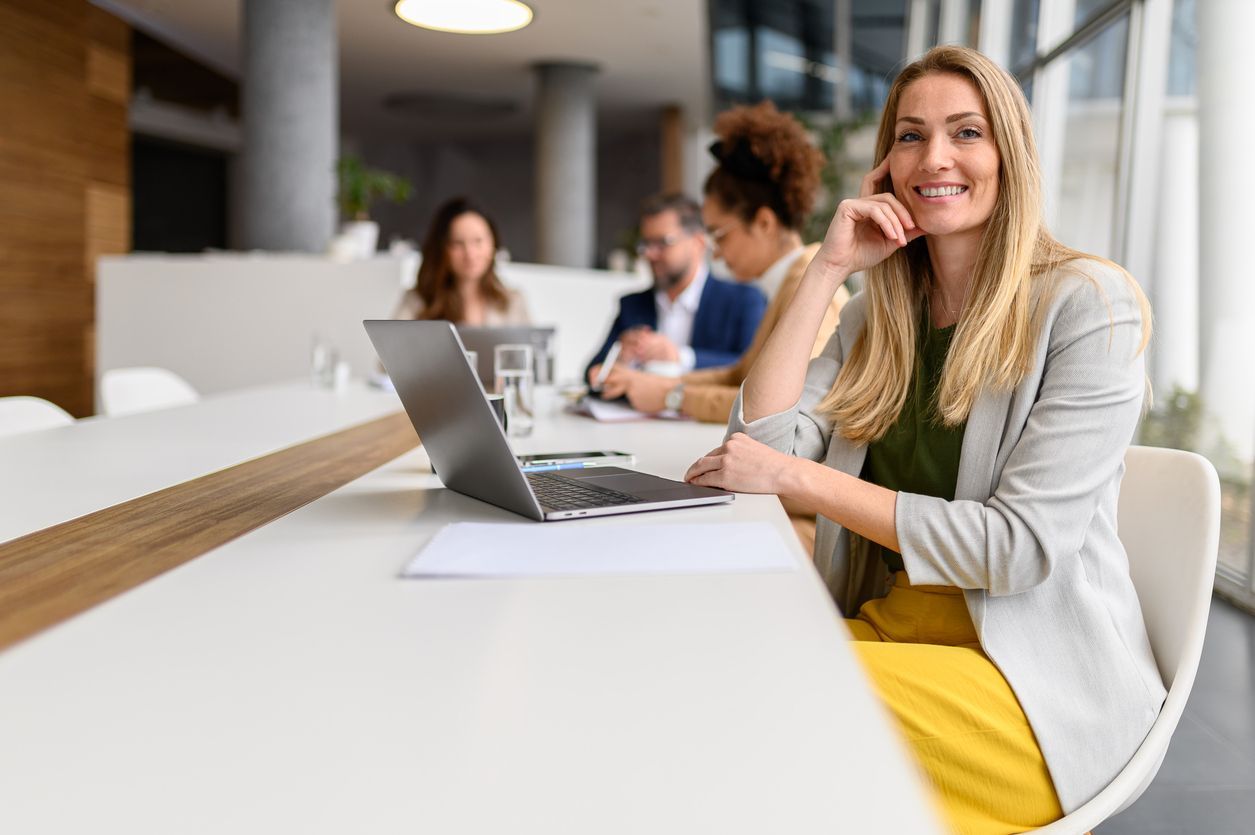 Woman smiling at a table with a laptop in a bright office lounge