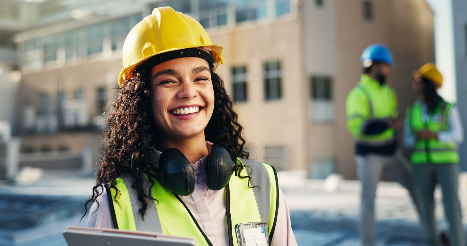 Smiling construction worker in yellow hard hat and vest at a worksite, with two workers blurred in the background