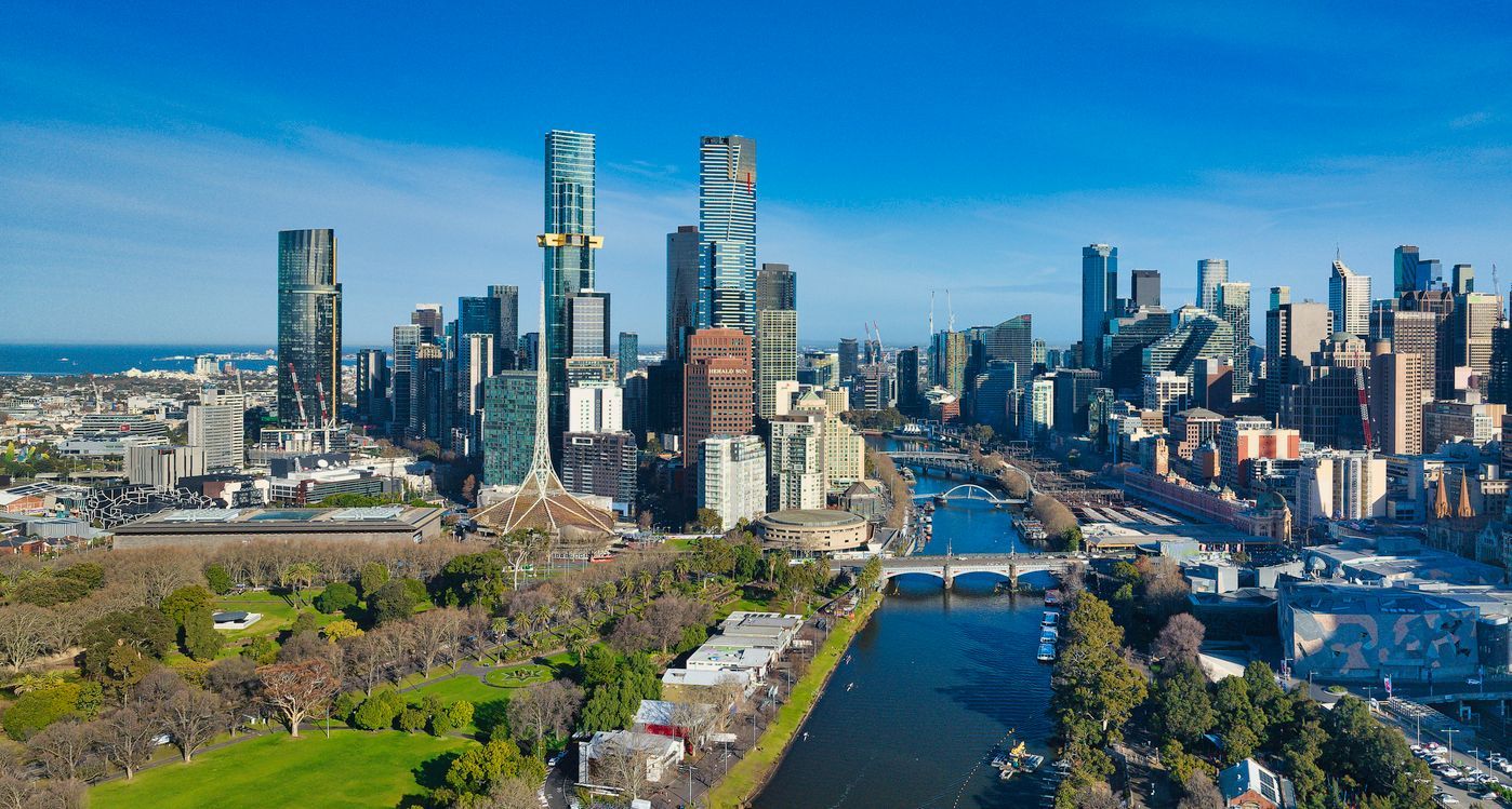 City skyline with tall modern buildings along a river and green parkland in the foreground.