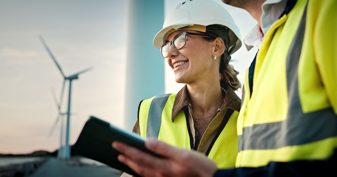 Two engineers in hard hats reviewing a tablet at a wind farm, with turbines in the background.