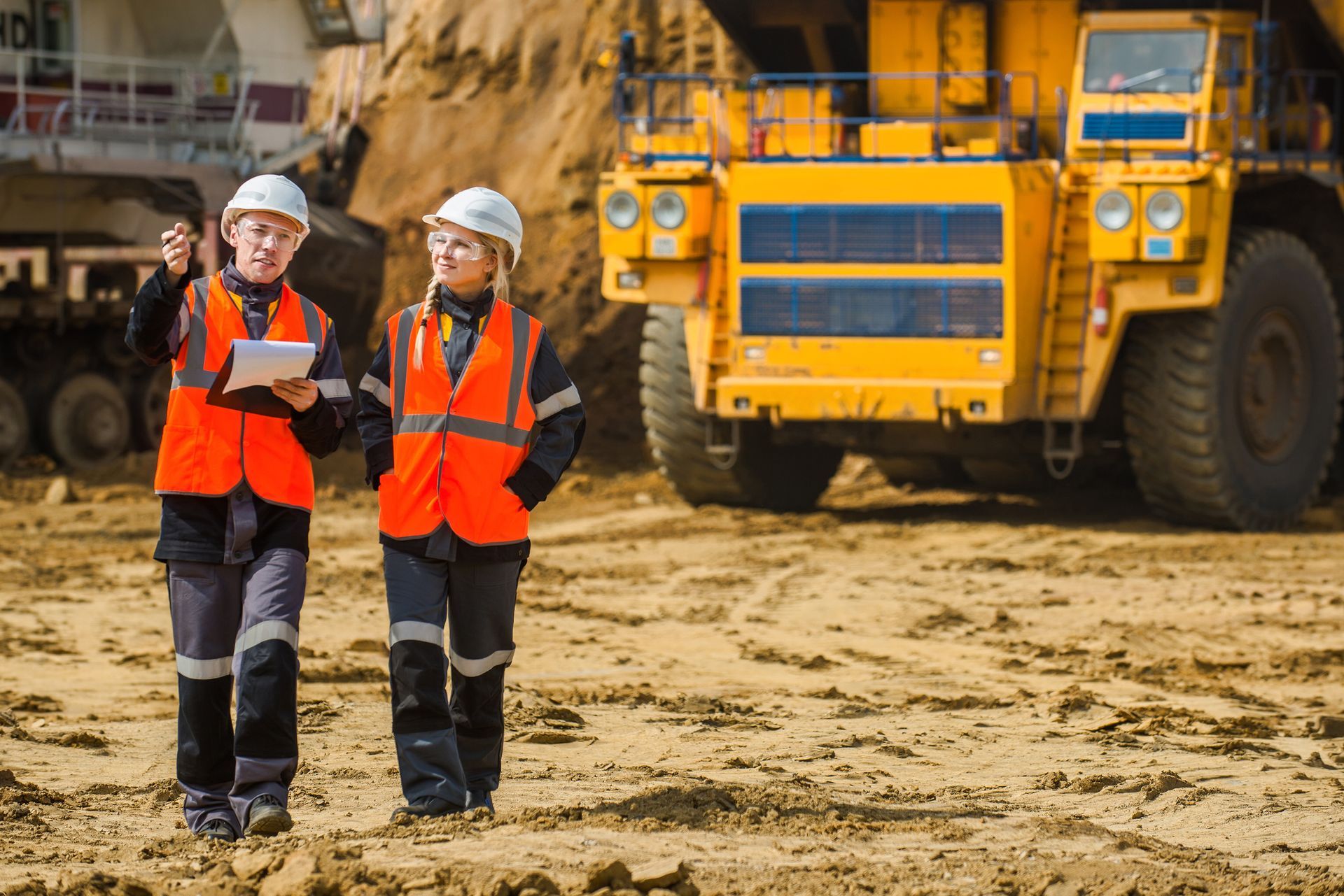 Two people in orange vests and hard hats at a mine, with large yellow truck.