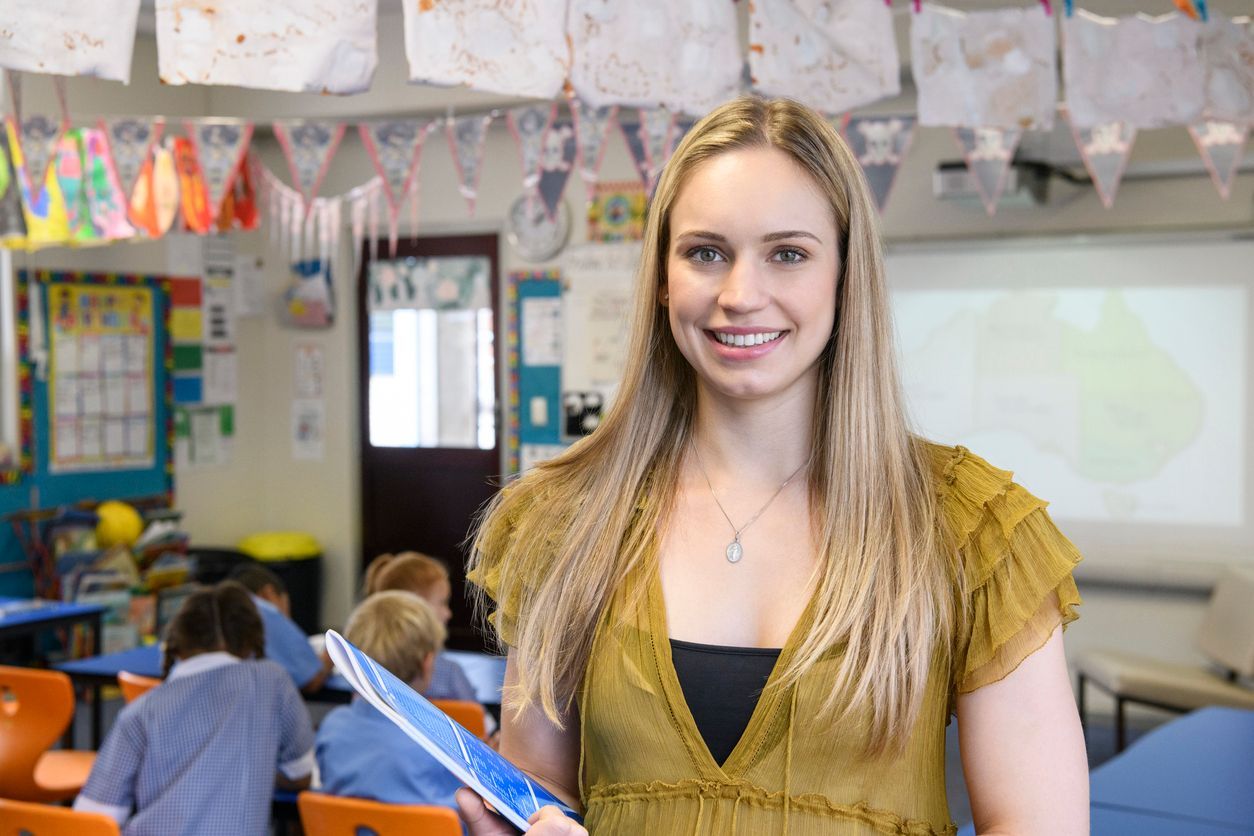 Teacher smiling in a classroom, holding a blue folder as students work in the background.
