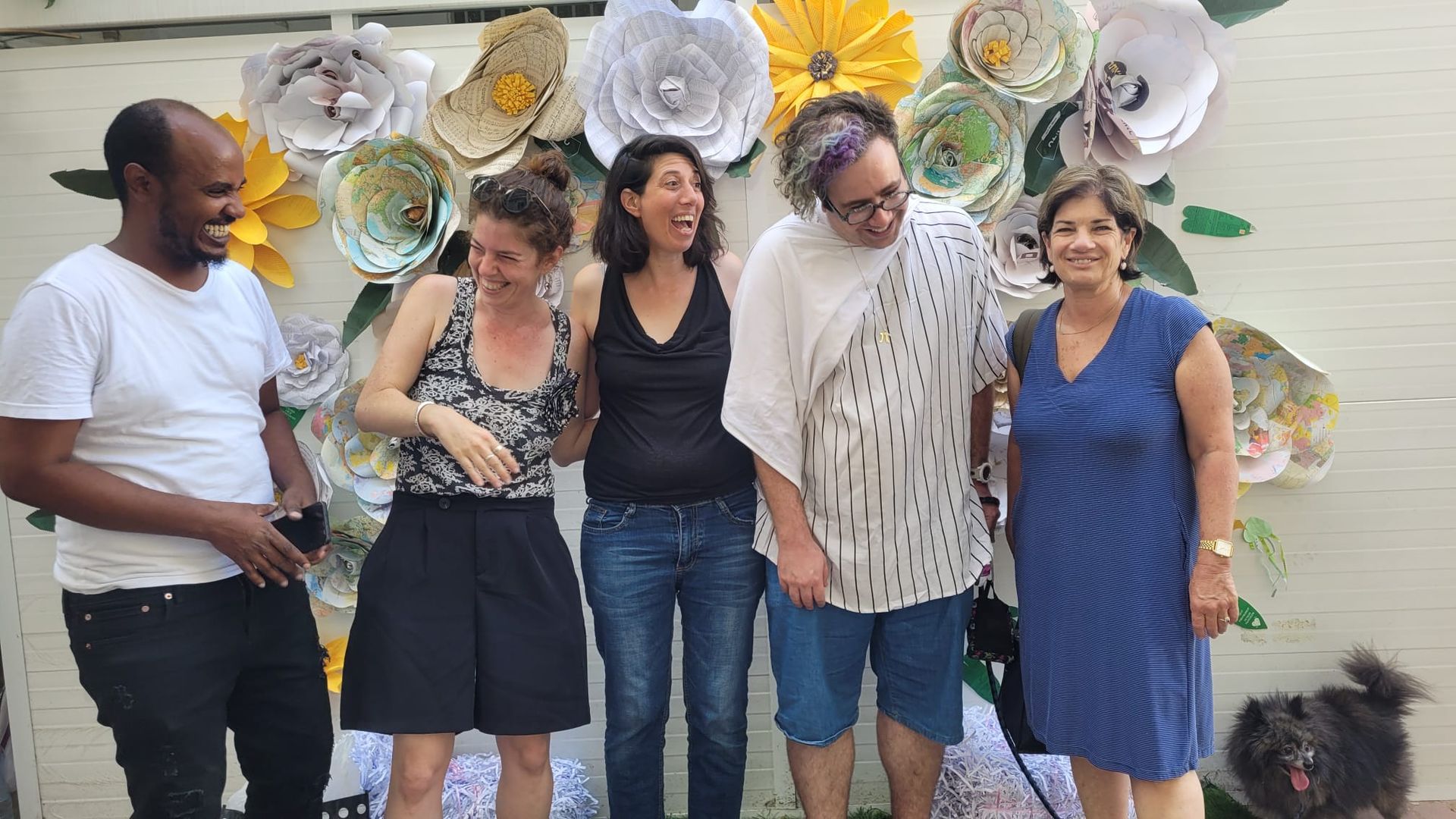 חמישה אנשים מצטלמים מול קישוטי פרחי נייר גדולים על קיר.