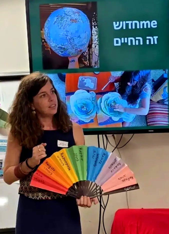 אישה מחזיקה מניפת קלפים צבעונית מול מסך המציג טקסט עברי וגרפיקה של כדור הארץ