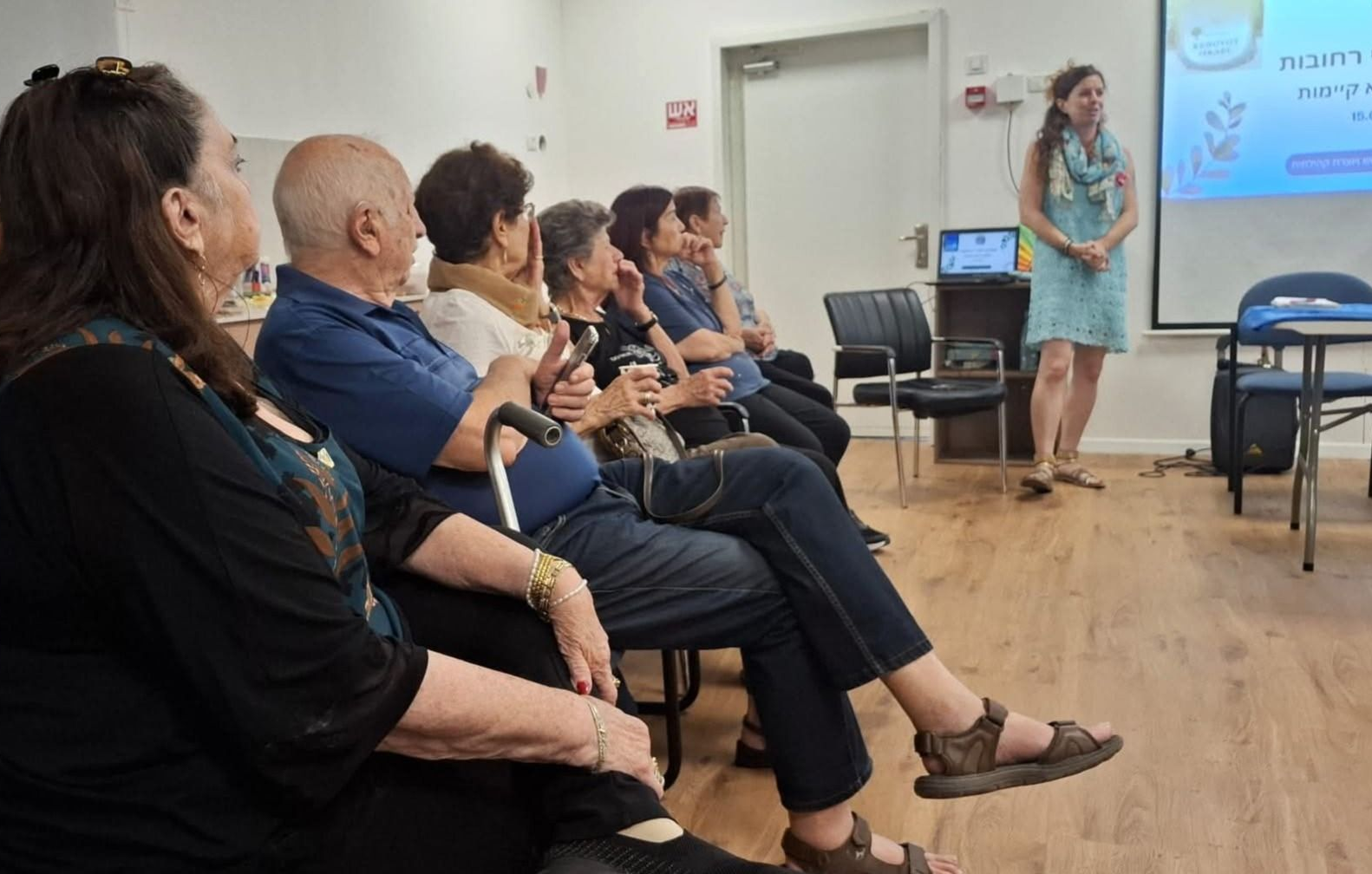 קהל יושב בחדר ישיבות וצופה במציג ליד שקופית מוקרנת