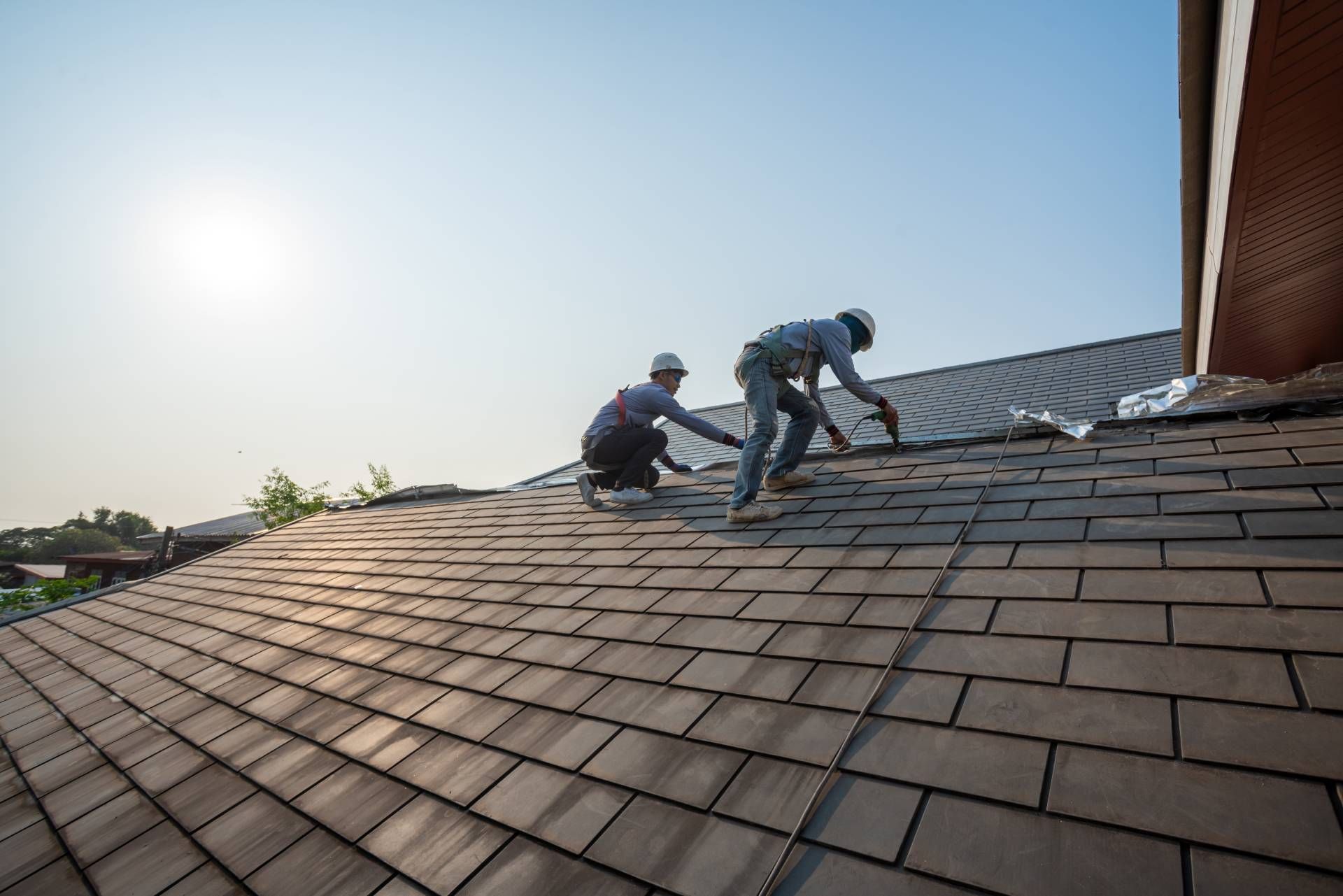 Two workers on a shingled roof during installation, with bright sunlight and a clear sky.