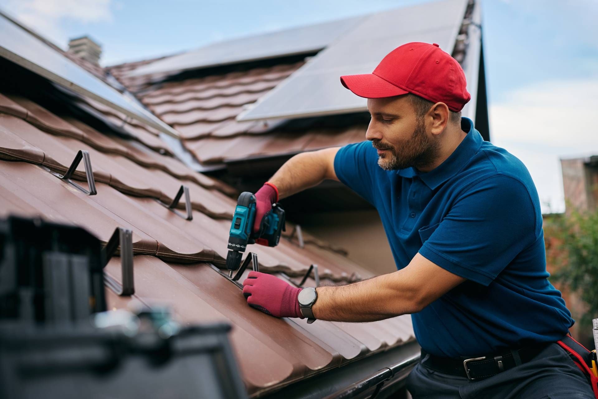 Worker in red cap and blue shirt fastening roof shingles with a drill