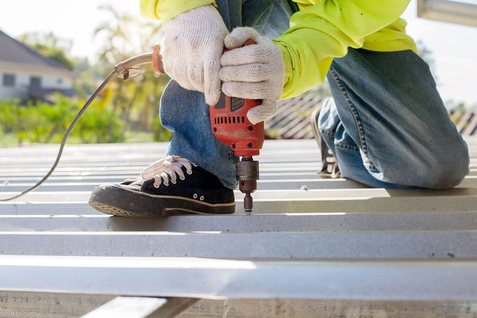 Worker drilling into a metal beam outdoors while kneeling in gloves and jeans.