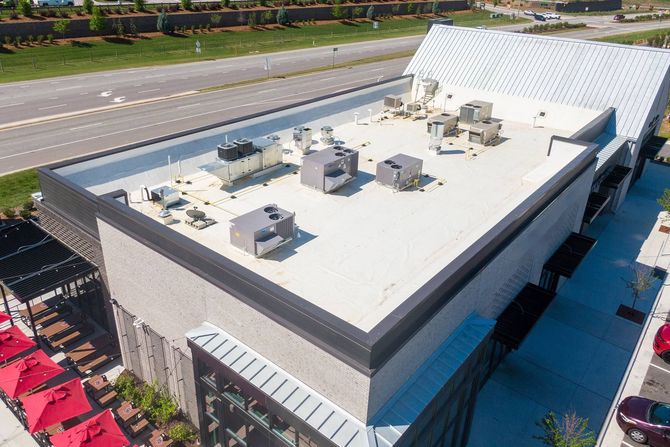 Aerial view of a flat-roofed building with HVAC units beside a highway and parking lot.