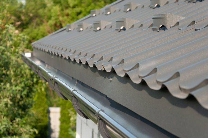 Gray corrugated metal roof with vents and gutters against a green tree background
