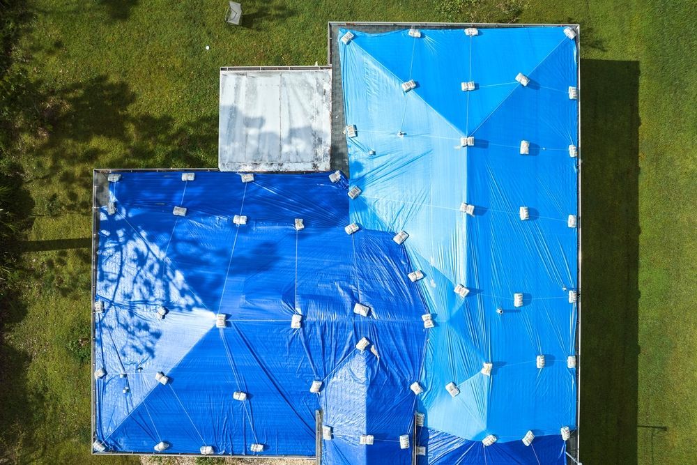Aerial view of a blue tarp-covered roof with a small white tarp on one side, surrounded by grass.