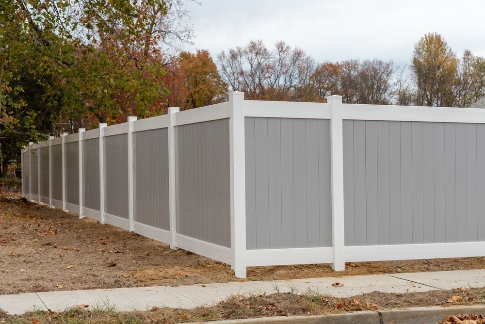 Gray privacy fence with white posts along a driveway, surrounded by trees in autumn