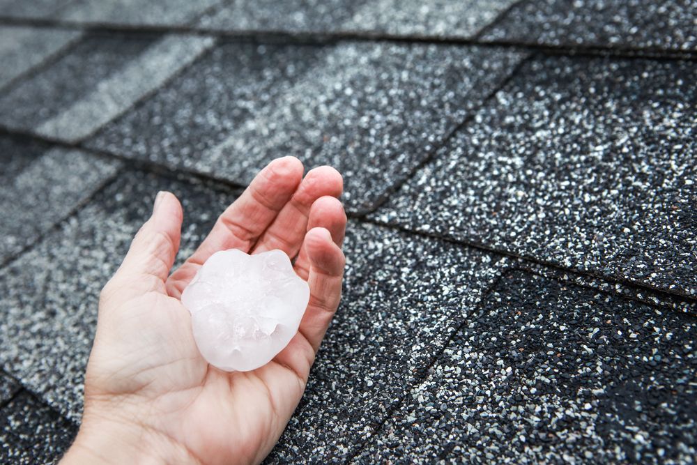 Hand holding a large hailstone over dark asphalt shingles
