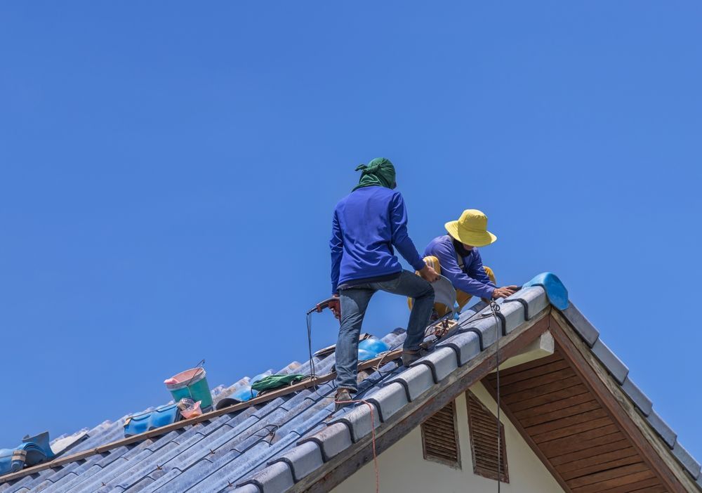 Two workers in blue and yellow gear repair a shingled roof under a clear blue sky.