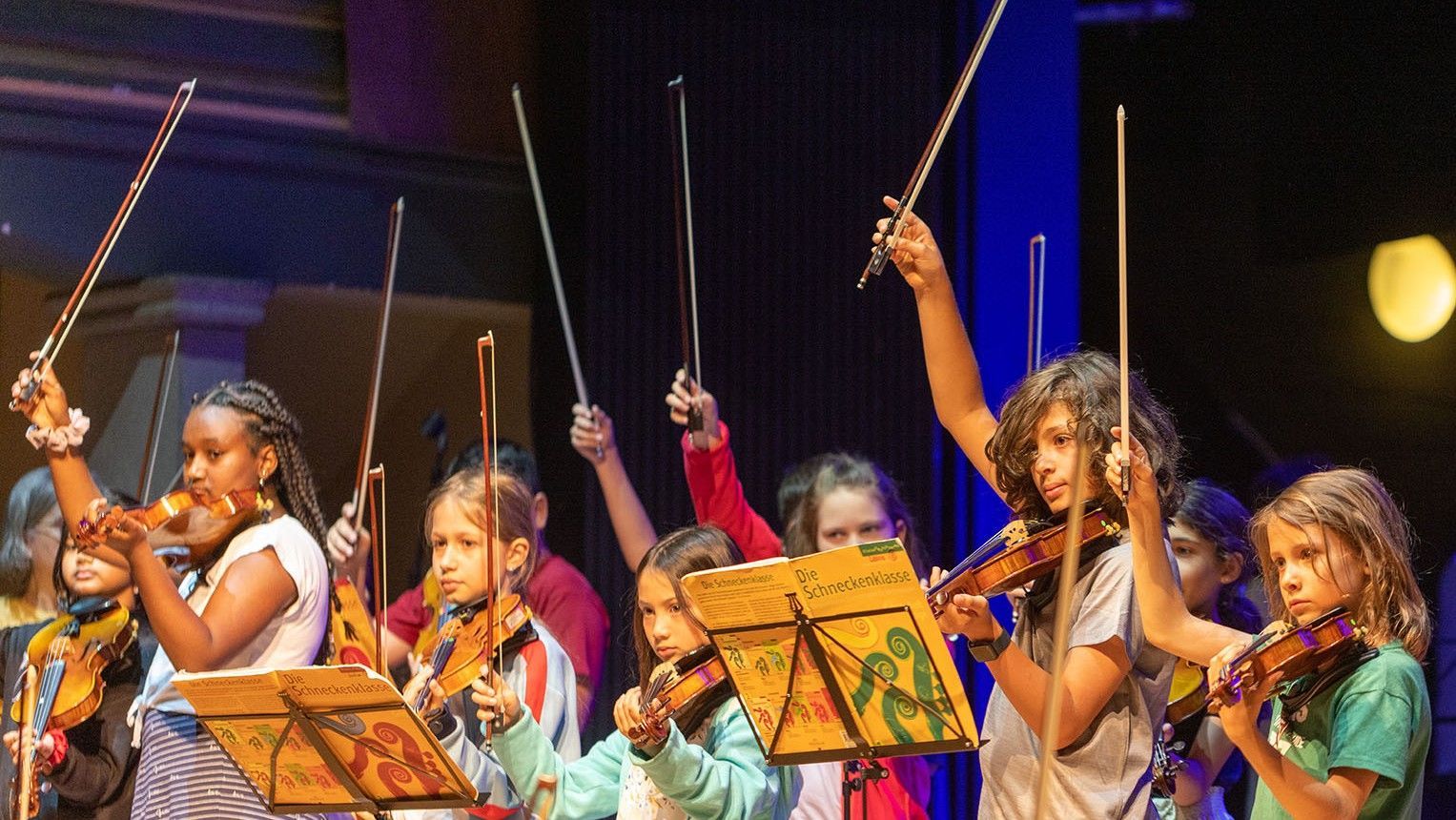 Musiker auf der Bühne, die Streichinstrumente spielen, die Bögen erhoben, vor ihnen Notenständer.