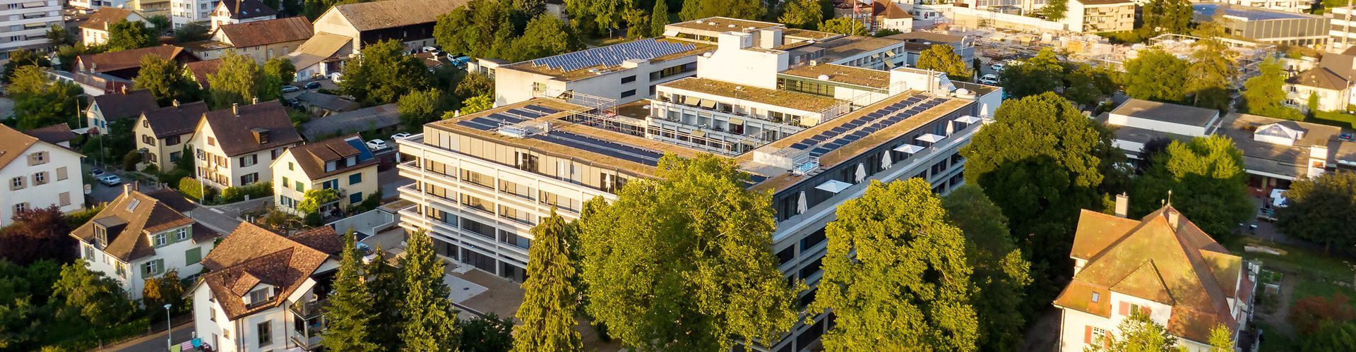 Luftaufnahme eines großen weißen Mehrfamilienhauses, umgeben von Bäumen und Wohnhäusern.