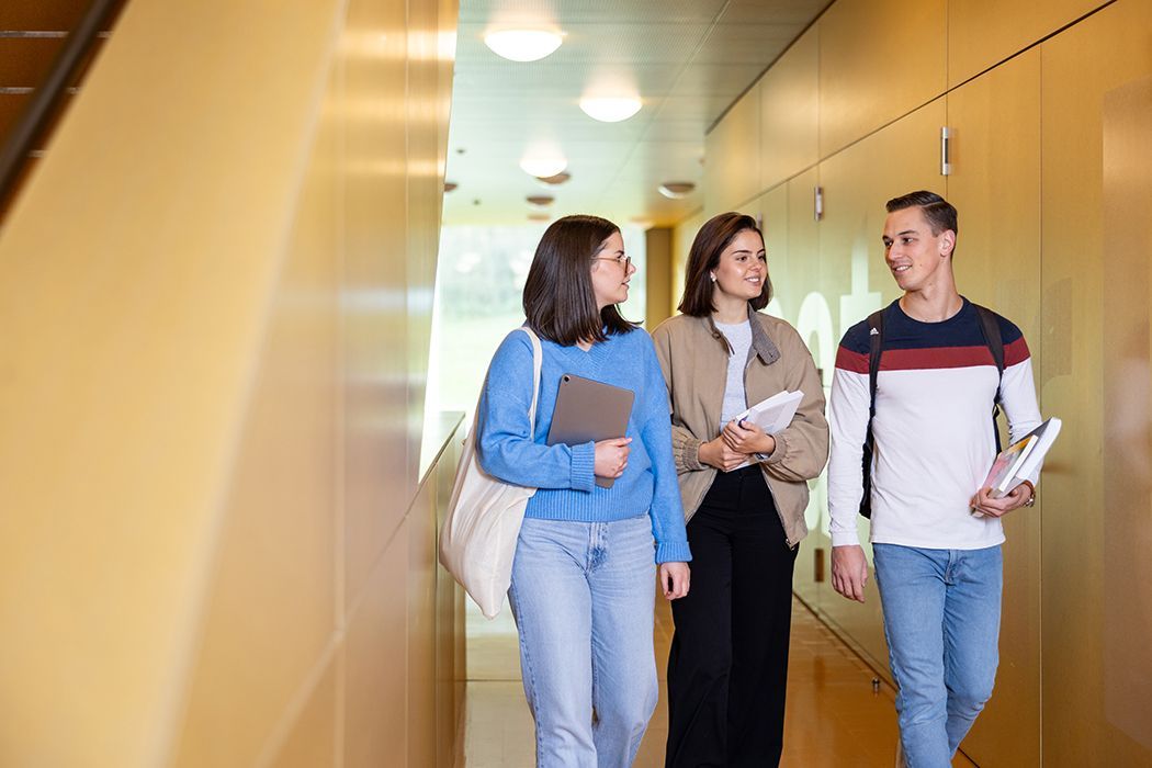 Drei Schüler gehen und unterhalten sich in einem hellen Flur; sie tragen Notizbücher und Ordner.
