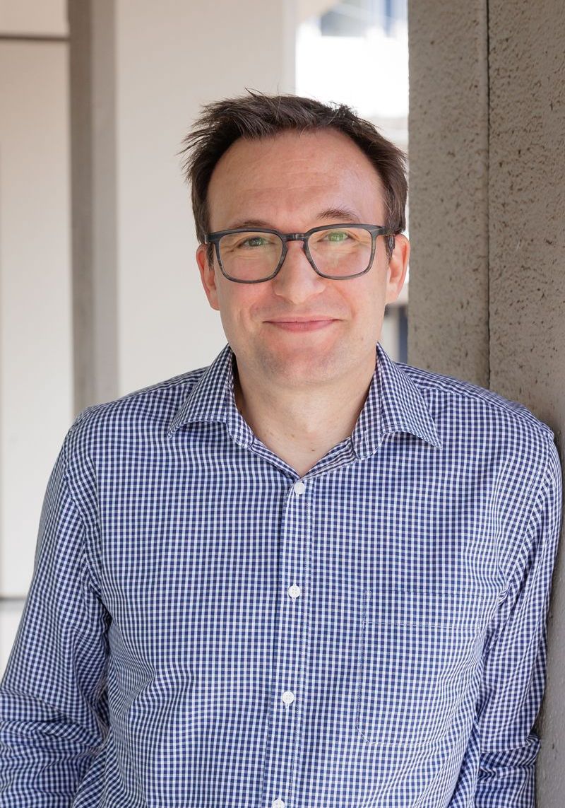 Ein Mann mit Brille und kariertem Hemd lehnt an einer Wand.