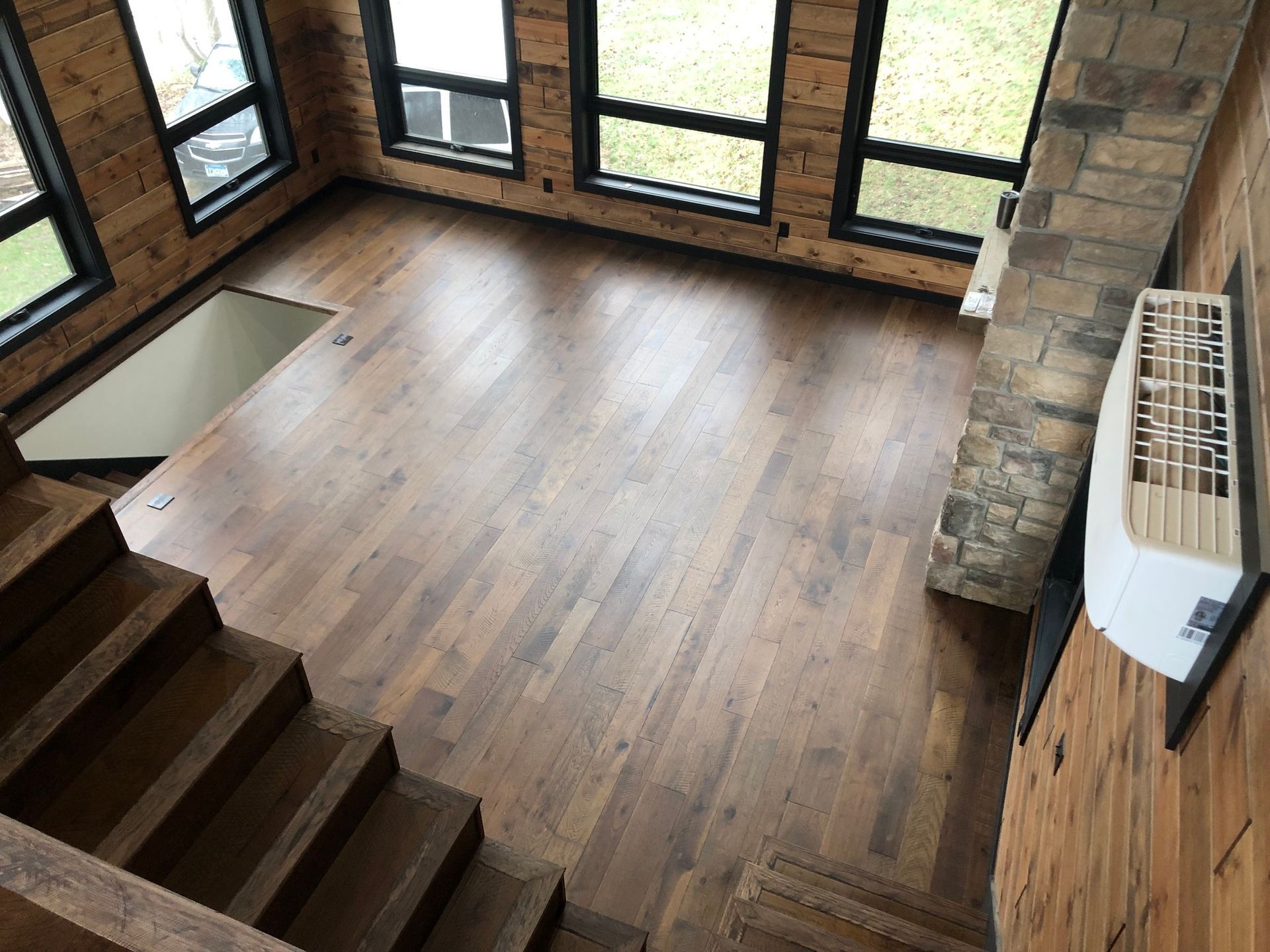 Empty wooden room with large windows, dark wood floors, and a stone fireplace.